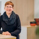 Meike Engling mit kurzen roten Haaren und Brille, trägt ein schwarzes Oberteil und gemusterten Schal, lächelt an einem Schreibtisch in einem hellen Büro.