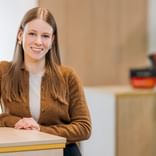 Porträt von Greta Martens mit langen braunen Haaren, in brauner Strickjacke und weißem Top, lächelnd an einem Schreibtisch in hellem Büro.