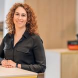 Irene Godoy Marin mit lockigem braunem Haar, in schwarzem Hemd und Uhr, lächelnd an einem weißen Tisch in einem modernen Büro.