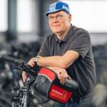 Robert Goldberg mit Brille und blauer Kappe, lehnt an Fahrrad mit roter Tasche im Fitnesscenter mit Heimtrainern im Hintergrund.