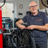 Herms Preuss mit Brille und grauem Poloshirt lächelnd in einer Fahrradwerkstatt, arbeitet an einem Fahrrad mit rotem Montageständer und Werkzeugen.