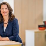Heike Salver with shoulder-length brown hair, wearing a navy blue blouse and necklace, sitting at a light-colored desk in a bright office.