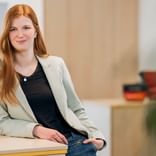 Phyllis Wiechmann with long red hair wearing a beige blazer and black top, standing in a modern office with blurred background.