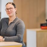 Pia Wilken mit Brille und zurückgebundenem Haar, trägt ein graues Langarmshirt und steht an einem Holztresen in einem hellen Büroraum.