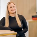 Cassandra Morla Silva mit langen blonden Haaren trägt ein schwarzes Langarmoberteil und steht an einem Empfangstresen in einem hellen Büroraum.