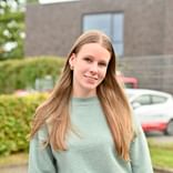 Greta Martens in mintgrünem Pullover draußen Porträt von Greta Martens mit langen blonden Haaren in einem mintgrünen Pullover, lächelt in die Kamera mit Backsteingebäude und Autos im Hintergrund.