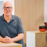 Markus Hüneke mit schwarzer Brille und grauem Poloshirt, sitzt an einem hellen Holztresen in einem hellen Büroraum.