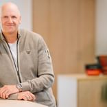 Andreas Bunge mit freundlichem Lächeln, trägt eine beige Strickjacke über einem weißen Hemd, steht in einem modernen Büroraum.