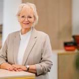 Anke Tammen mit kurzen blonden Haaren und Brille, trägt einen beigen Blazer über einem weißen Rollkragenpullover, steht in einem hellen Büro.