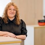 Marscha Büsing mit lockigem blondem Haar in schwarzer Bluse, sitzt an einem hellen Holztisch in einem hellen Büro mit unscharfem Hintergrund.
