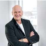 Andreas Bunge mit verschränkten Armen, trägt einen dunklen Blazer über einem weißen Hemd, lächelt in einem hellen Büro mit Fenstern im Hintergrund.