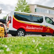 Weiß-roter Rückenwind Transporter auf Wiese geparkt mit Koffern und gelbem Rucksack daneben. Wohngebäude im Hintergrund sichtbar.