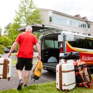 Mann im roten Shirt lädt weiße Koffer in einen Rückenwind-Transporter mit offenen Hecktüren in Wohngebiet.