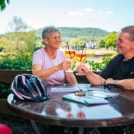 Zwei Radfahrer stoßen mit Weingläsern am Außentisch in Wehrden an. Fahrradhelme auf dem Tisch, Fluss und Hügel im Hintergrund.