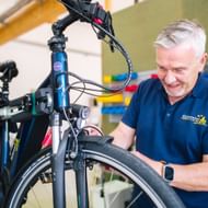 Älterer Mann mit grauen Haaren in blauem Poloshirt arbeitet an schwarzem Fahrrad in professioneller Reparaturwerkstatt mit Werkzeugen.