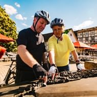 Zwei Radfahrer mit Helmen studieren eine Karte an einem Holztisch auf Melsungens historischem Marktplatz mit Fachwerkhäusern im Hintergrund.