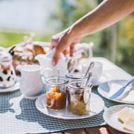 Frühstückstisch im Freien mit weißer Teekanne, Marmeladengläsern, Butterteller und Blumentopf auf karierter Tischdecke.