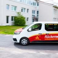 Weißer und roter Rückenwind-Van mit Fahrrad-Logo fährt auf einer Straße an weißen Gebäuden und grünen Bäumen vorbei in Bewegungsunschärfe.