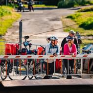 Zwei Radfahrer mit Helmen und Fahrrädern stehen auf einer kleinen Fährplattform in Hemeln. Die Fähre hat rote Ausrüstung und überquert einen Fluss.