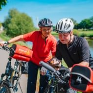 Zwei Radfahrer mit Helmen stehen mit ihren E-Bikes auf einem Weg neben der Weser, umgeben von grünen Bäumen unter blauem Himmel.