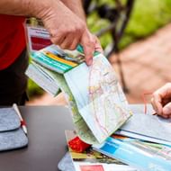 Zwei Personen betrachten Reisebroschüren und Karten auf einem Tisch. Eine Person im roten Shirt hält bunte Reiseunterlagen.
