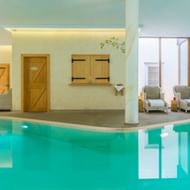 Modernes Hallenbad mit türkisfarbenem Wasser, weißen Säulen, Holztüren und gemütlichem Sitzbereich mit grauen Sesseln.