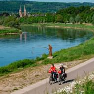 Zwei Radfahrer auf befestigtem Weg entlang der Weser mit Angler am Flussufer. Historisches Höxter mit Doppeltürmen im Hintergrund.