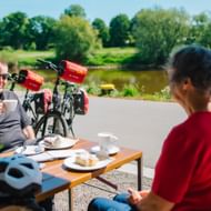 Zwei Radfahrer genießen Kaffee an Außentisch nahe der Weser in Höxter. Fahrräder mit roten Taschen dahinter, grüne Landschaft sichtbar.