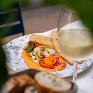 Dekorativer Teller mit Hummus, geschnittenem Gemüse, Kräutern und Brot, serviert mit einem Glas Weißwein auf einem Tisch im Freien.