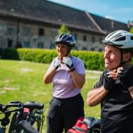 Zwei Radfahrer mit Helmen machen Pause am Kloster Corvey. Historische Steingebäude und Kirchtürme im Hintergrund mit grüner Wiese.