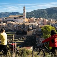 Zwei Radfahrer mit Rädern betrachten ein Dorf am Hang mit Kirchturm in der Region Valencia, Spanien. Berge erheben sich im Hintergrund.