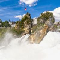 Mächtiger Rheinfall mit weißem Wasser, das über Felsen stürzt. Eine Schweizer Fahne weht von einem Felsvorsprung in der Mitte, Gebäude im Hintergrund.