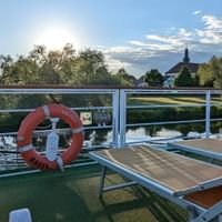 Sonnendeck der MS Casanova mit orangen und beigen Liegestühlen, weißen Geländern, Rettungsring und Flussblick mit Kirchturm im Hintergrund.