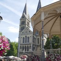 Gotische Kirche mit Zwillingstürmen hinter einem verzierten weißen Pavillon mit rosa und weißen Blumen. Historische Laterne und blühende Bäume sichtbar.