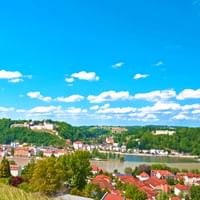 Panoramablick auf Passau mit historischen Gebäuden und roten Ziegeldächern entlang der Donau, umgeben von grünen Hügeln unter blauem Himmel.