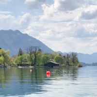 Ruhiger See im Salzkammergut mit roten Bojen, Ufergebäuden und Bergen unter bewölktem Himmel. Bäume säumen das Ufer.