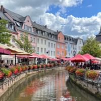 Historisches Stadtzentrum von Saarburg mit bunten Fachwerkhäusern entlang eines Kanals. Rote Sonnenschirme beschatten Außengastronomie.
