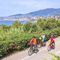 Drei Radfahrer in bunten Trikots fahren auf einer Küstenstraße mit Mittelmeer und Sanremo-Stadtbild im Hintergrund.
