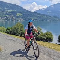 Radfahrerin mit blauem Helm fährt auf befestigtem Weg entlang großem Alpensee mit bewaldeten Bergen und Dörfern im Hintergrund.