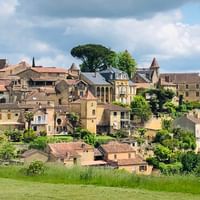 Dorf am Hang in der Dordogne mit Steingebäuden, Kirchturm und Terrakottadächern, umgeben von grünen Feldern und Bäumen unter bewölktem Himmel.