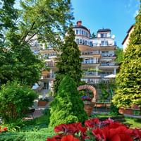 Mehrstöckiges Hotelgebäude mit Balkonen, umgeben von üppigen grünen Gärten mit roten Blumen, alten Bäumen und gepflegten Anlagen.