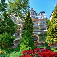 Mehrstöckiges Hotel in Masuren mit Balkonen, umgeben von üppigem Garten mit hohen Bäumen, geformten Koniferen und roten Blumen im Vordergrund.