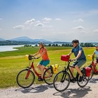 Drei Radfahrer mit Tourenrädern und Packtaschen auf Schotterweg durch grüne Felder. See und Berge im Hintergrund unter blauem Himmel.