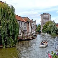 Touristenboot auf Kanal in Brügge mit historischen Gebäuden, Trauerweide und modernem Turm im Hintergrund. Bunte Blumen im Vordergrund sichtbar.