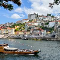 Traditionelles Holzboot mit Passagieren auf dem Douro in Porto, mit bunten historischen Gebäuden am Hang und der Dom Luís I Brücke.
