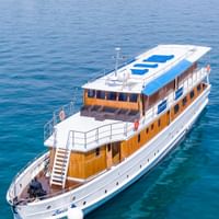 Luftaufnahme der MY Linda, ein weißes Kreuzfahrtschiff mit Holzdeck und blauen Sonnenliegen auf dem Dach, vor Anker in türkisfarbenem Wasser.