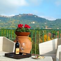 Außenterrasse mit Korbstühlen, Weinflasche und Glas auf Tablett, rote Blumen in Terrakotta-Topf. Grüne Hügel mit Bergdörfern im Hintergrund.