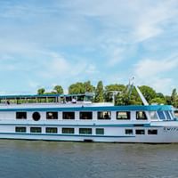 Weißes Flusskreuzfahrtschiff MS Switzerland mit blauem Zierstreifen auf ruhigem Wasser. Bäume und blauer Himmel im Hintergrund.