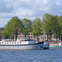 Weiß-blaues Hausboot Floor van Aemstel im Hafen mit Bäumen und historischen Gebäuden am Ufer unter bewölktem Himmel.