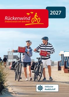 Titelbild des Rückenwind Radreisen Katalogs 2027 zeigt zwei Radfahrer mit Helmen, die Fahrräder auf einer Strandpromenade schieben.